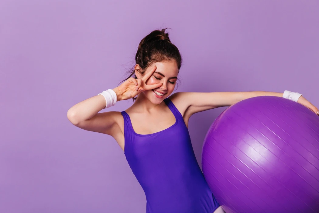 Slim gymnast woman holds fitball and shows peace sign on purple wall